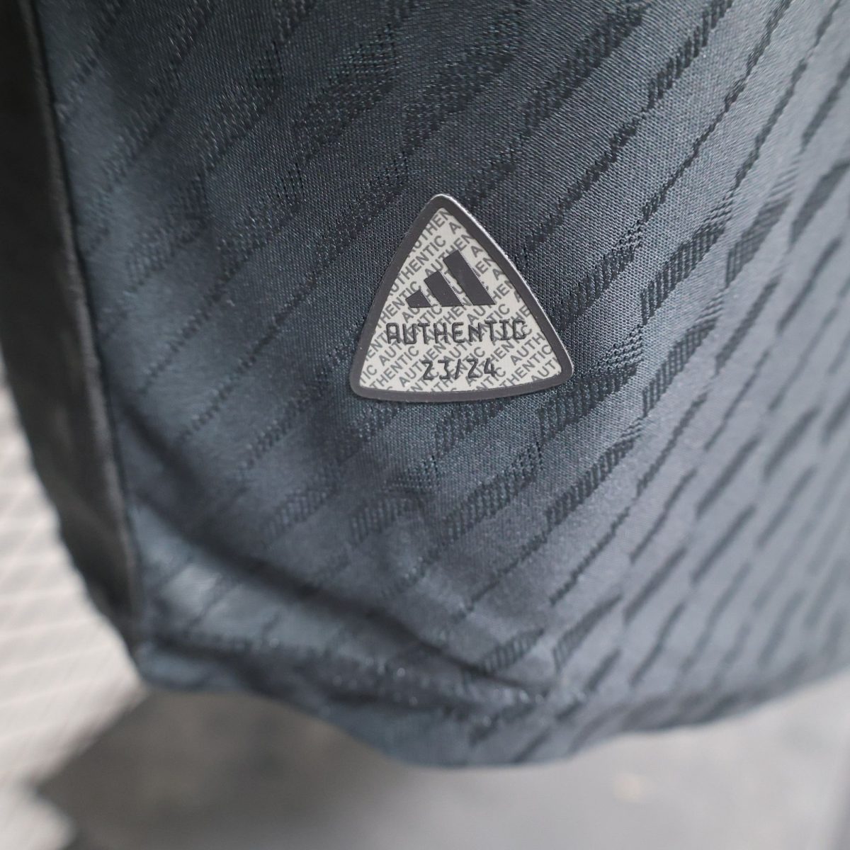 Close-up of a triangular badge/logo on dark fabric, with the word 'AUTHENTIC' repeated in a pattern across the surface.