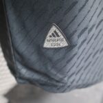 Close-up of a triangular badge/logo on dark fabric, with the word 'AUTHENTIC' repeated in a pattern across the surface.