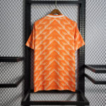 Orange short-sleeve shirt with a white geometric chevron pattern on a hanger against a metal display rack.