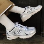 f2087cea-ea34-4b70-8556-ed679a2f2b0c | UAE SPORT STORE Person wearing white socks and silver-white New Balance sneakers resting on a dark doorstep or threshold.