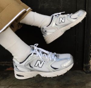 f2087cea-ea34-4b70-8556-ed679a2f2b0c | UAE SPORT STORE Person wearing white socks and silver-white New Balance sneakers resting on a dark doorstep or threshold.