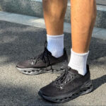 Person wearing black athletic sneakers and white crew socks standing on a paved surface near concrete steps.