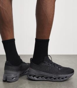 Person wearing black athletic shoes with thick, hollow-cut sole and black socks, standing on a gray floor.