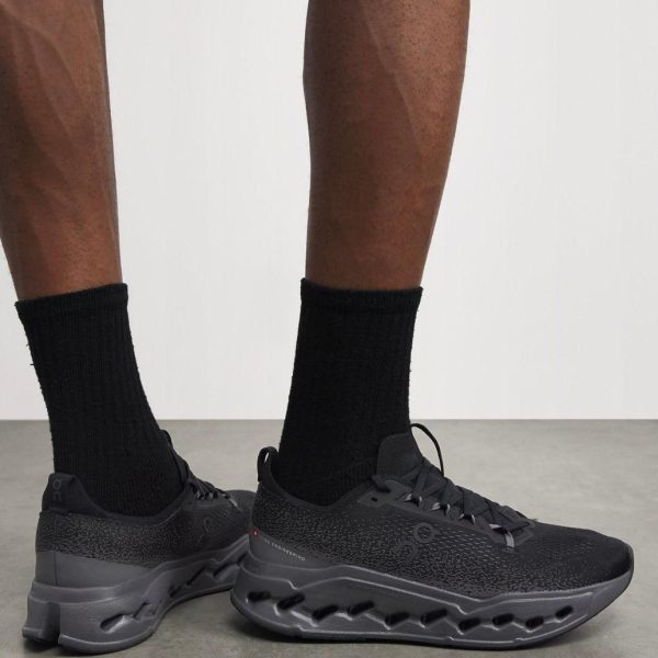 Person wearing black athletic shoes with thick, hollow-cut sole and black socks, standing on a gray floor.