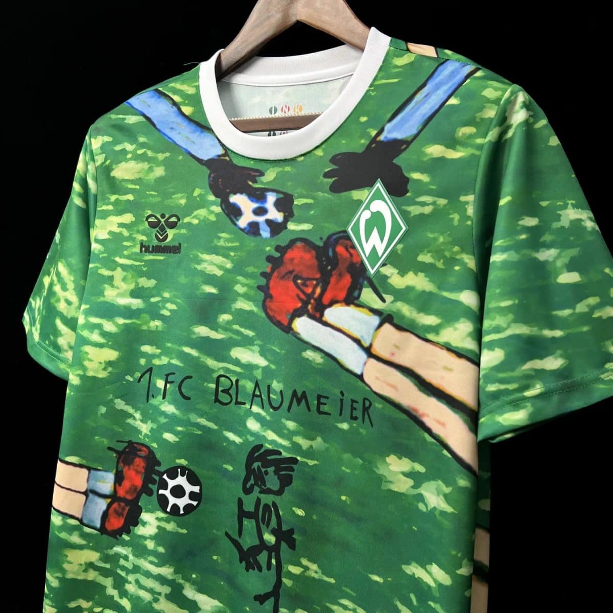 Green sports jersey on a hanger with a cartoon hand graphic, Werder Bremen crest, and Hummel logo; handwritten '1.FC BLAUMEIER' text visible.