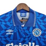 Blue Napoli football jersey on a hanger, featuring the Umbro logo and the Napoli crest, with a geometric dark-blue pattern across the fabric.