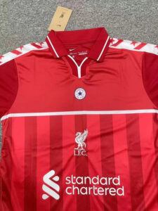 Red Liverpool FC jersey laid flat showing crest, sponsor logo 'standard chartered', and white-accented collar; tag tucked near neck.