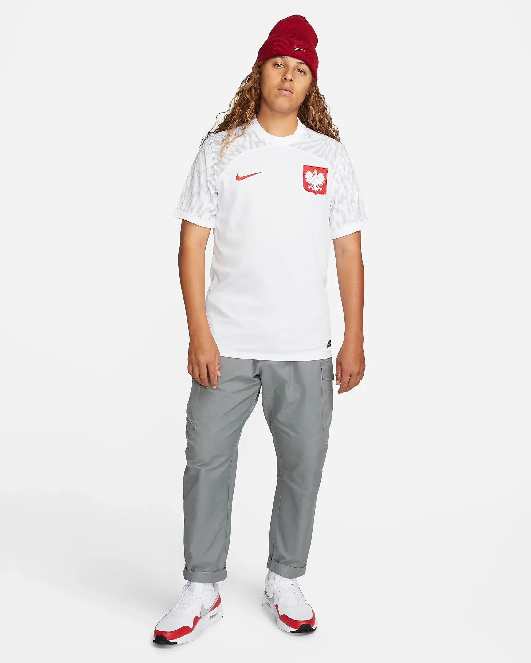 f32113f9-f30c-45ab-84fe-1a02e24b4235_cb63c7e5-3d4a-4db2-b590-e6ead9e47139 | UAE SPORT STORE Full-body shot of a person wearing a white athletic shirt with a red Polish eagle crest, gray cargo pants, white sneakers, and a red beanie.