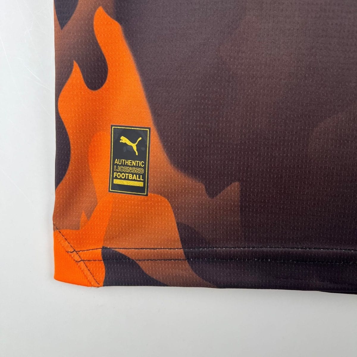 Close-up of orange and black football jersey fabric with a small Puma logo badge reading "AUTHENTIC LICENSED FOOTBALL"