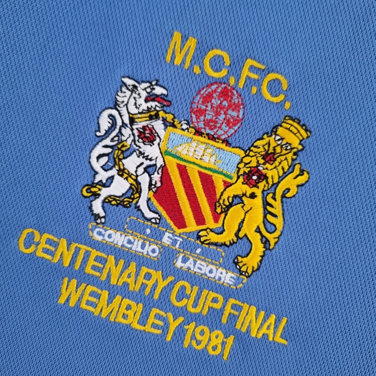 Close-up of blue fabric with a yellow-embroidered crest and text at the bottom, showing detailed stitching of a heraldic emblem.