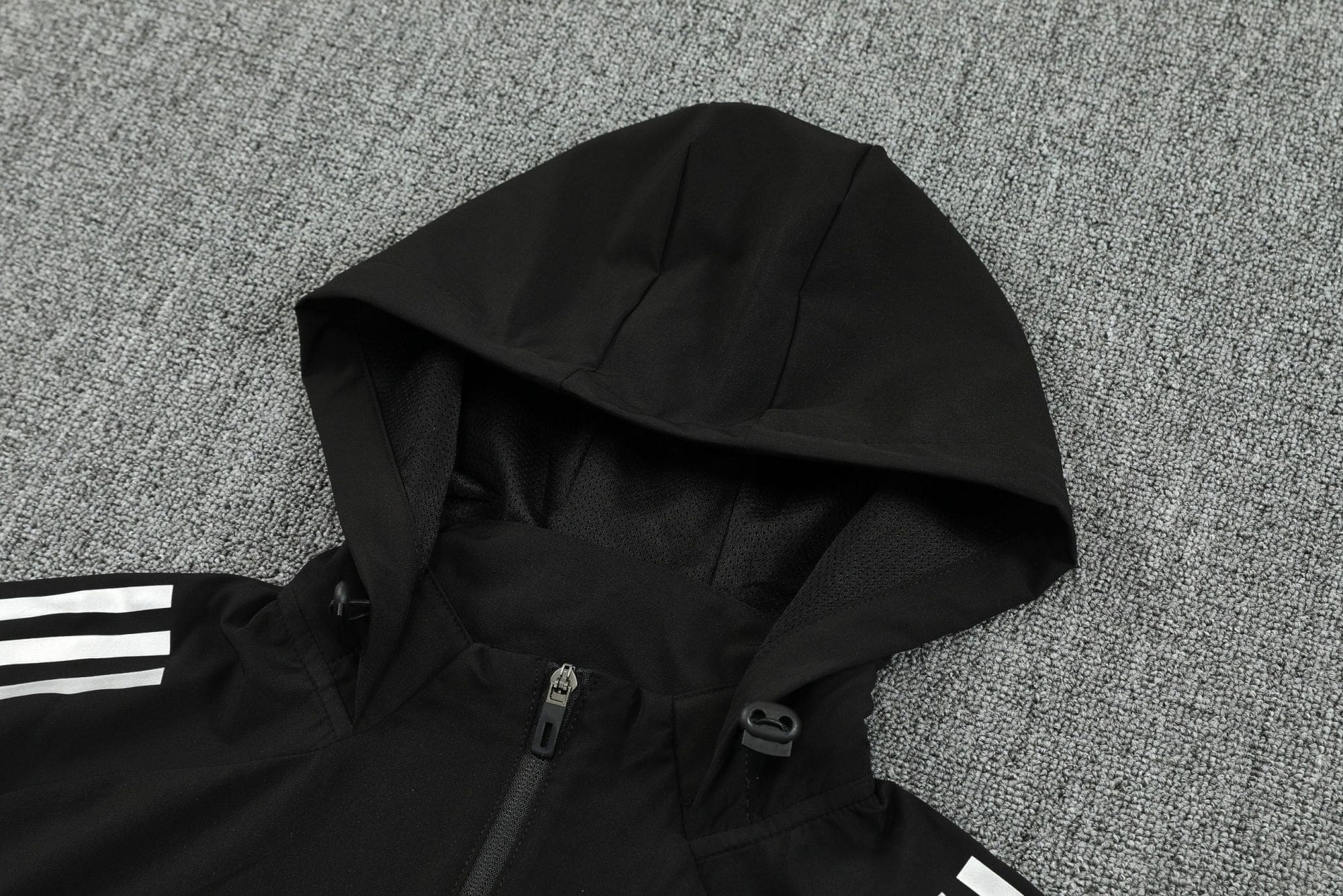 f44dc1ca | UAE SPORT STORE Black hooded jacket laid flat on a gray carpet, zipper visible along the front, white striped sleeve detail partially shown.