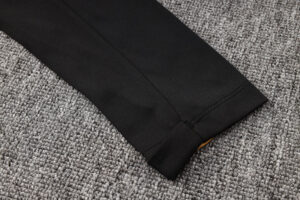 Black pant leg cuff resting on a gray textured carpet, with a small yellow tag show­ing at the seam edge.