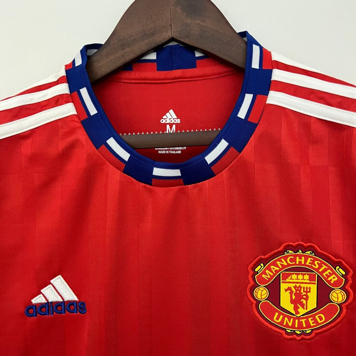 Red Manchester United jersey on a wooden hanger, Adidas logo visible at collar, crest partly shown on right side.
