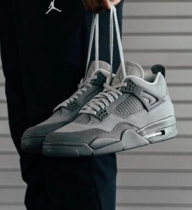 Gray athletic sneakers are being held by their laces in front of a dark outfit and a gray siding background.