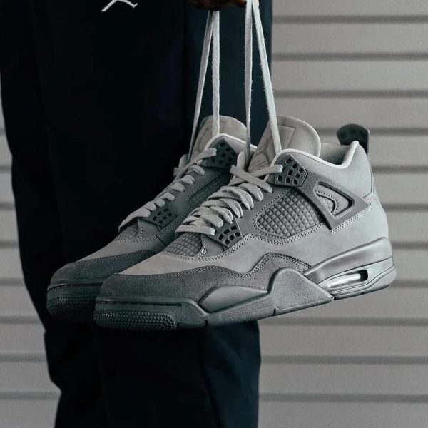 Gray athletic sneakers are being held by their laces in front of a dark outfit and a gray siding background.