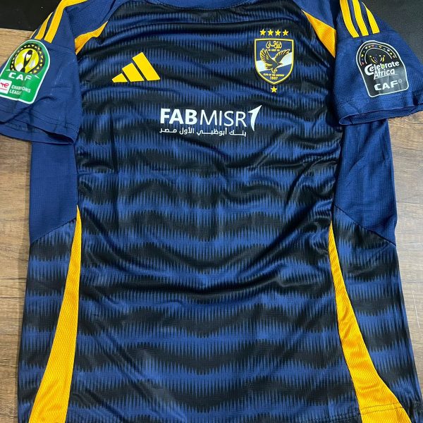 Dark blue zigzag-patterned soccer jersey with yellow side panels and stripes, crest on the left chest, Adidas logo, and sponsor text FAB MISR across the midsection, worn look suggesting a game jersey and CAF patches on the sleeves.