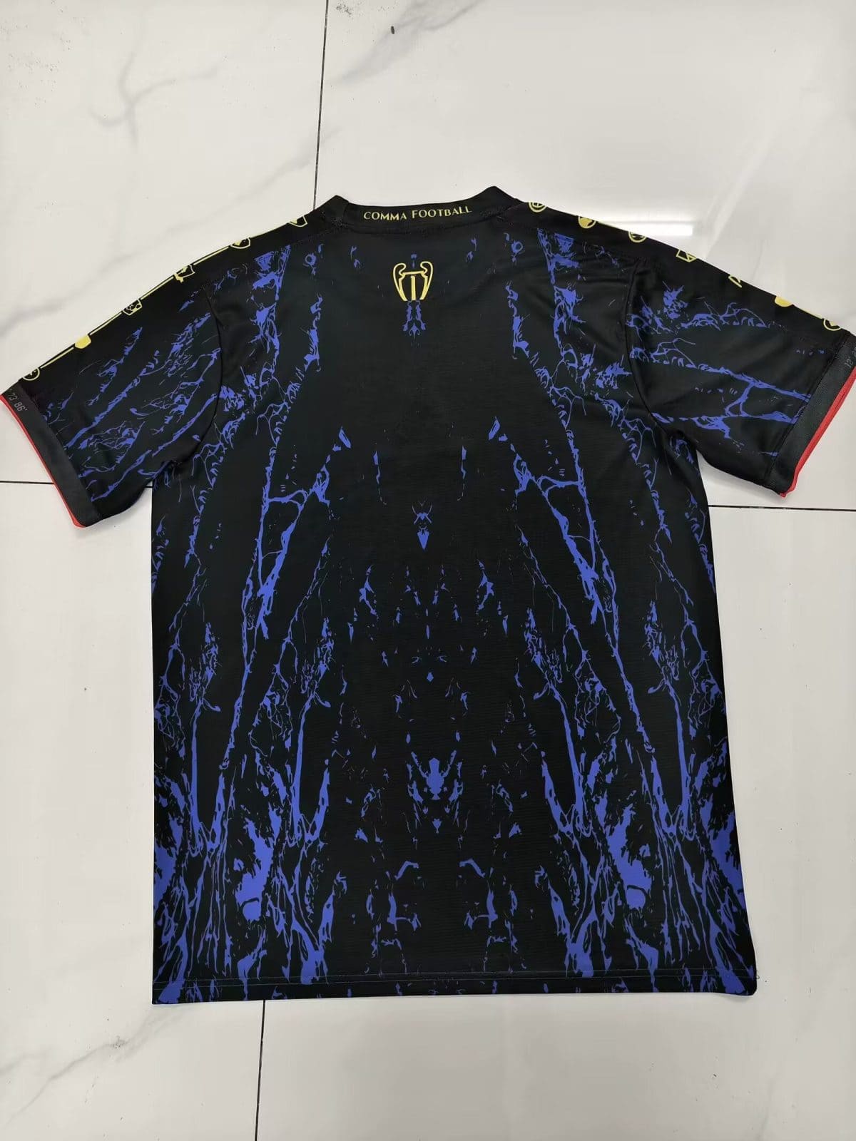 Black football jersey with a blue marbled pattern, small gold trophy emblem at the upper back, and 'COMMA FOOTBALL' on the collar with red-trimmed sleeves.