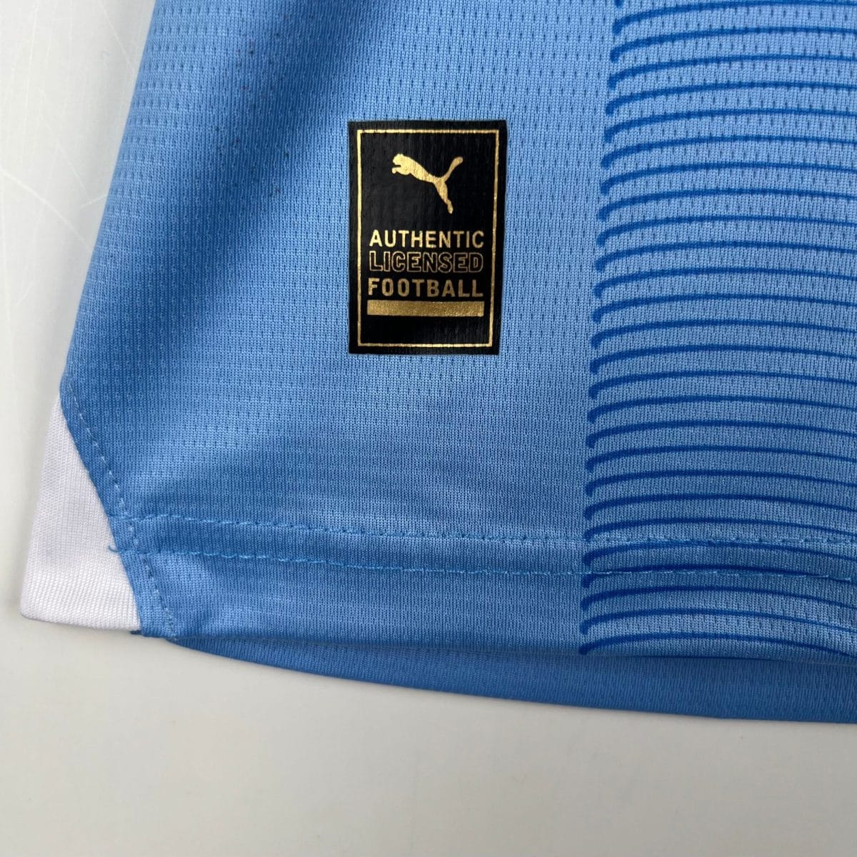Close-up of blue sports jersey fabric with a black and gold patch showing the Puma logo and text 'AUTHENTIC LICENSED FOOTBALL'