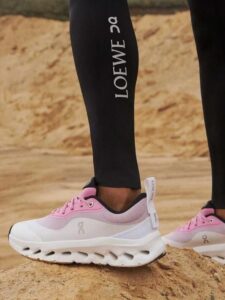 f7b58457-54a4-4050-a126-44ff66aeb31b | UAE SPORT STORE Pink-and-white athletic sneakers on a person standing on sand, LOEWE leggings visible.