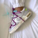 White low-top sneaker with bright purple laces and teal tag, resting on a white bedsheet with a cuffed pant leg visible.