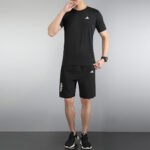 Man in a black Adidas athletic outfit (tee, shorts, sneakers) standing against a gray backdrop, posing with a hand near the mouth.