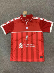 Red Liverpool FC jersey with vertical darker red stripes, white sponsor logo 'standard chartered' across the front, club crest and bird emblem, white-trimmed collar, and white sleeve cuffs hanging on a gray carpet.