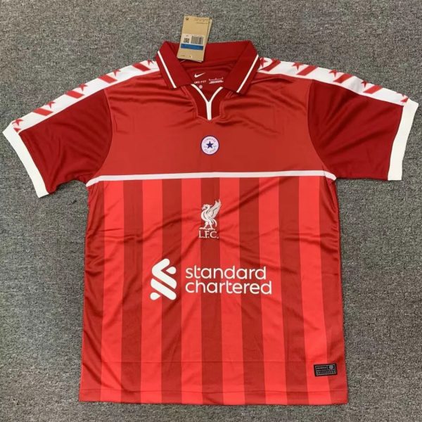 Red Liverpool FC jersey with vertical darker red stripes, white sponsor logo 'standard chartered' across the front, club crest and bird emblem, white-trimmed collar, and white sleeve cuffs hanging on a gray carpet.