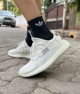 White knit sneakers with laces being worn with black Adidas socks on a cobblestone street, mid-step in an urban setting.