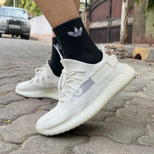 White knit sneakers with laces being worn with black Adidas socks on a cobblestone street, mid-step in an urban setting.