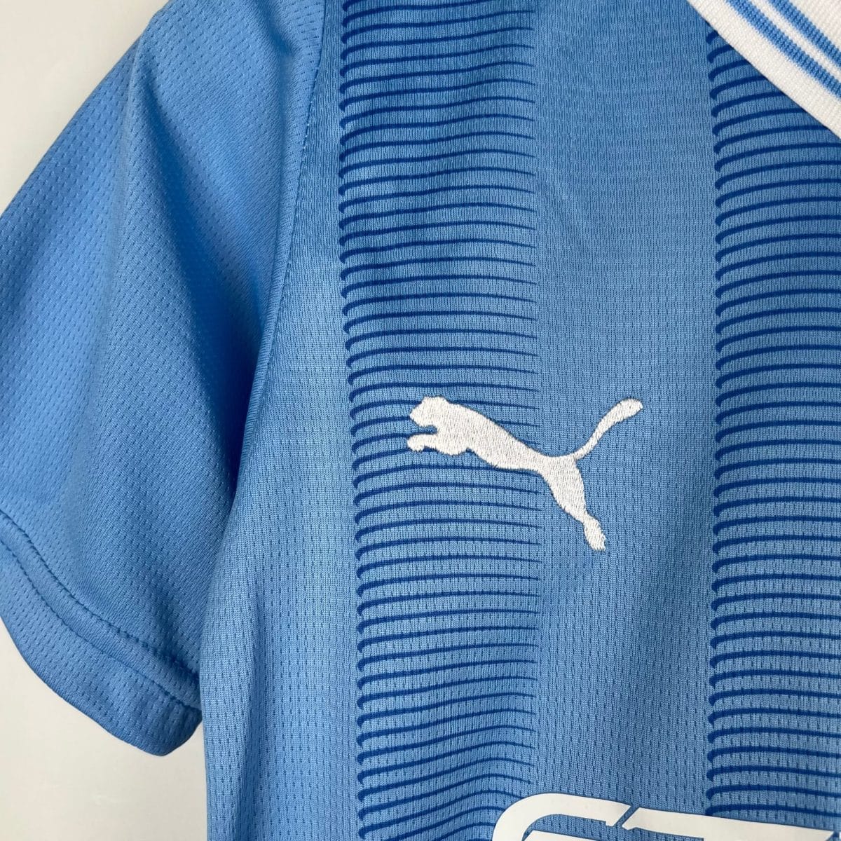 Close-up of blue athletic jersey fabric with small perforations and a partial white logo/lettering visible at the bottom-right.