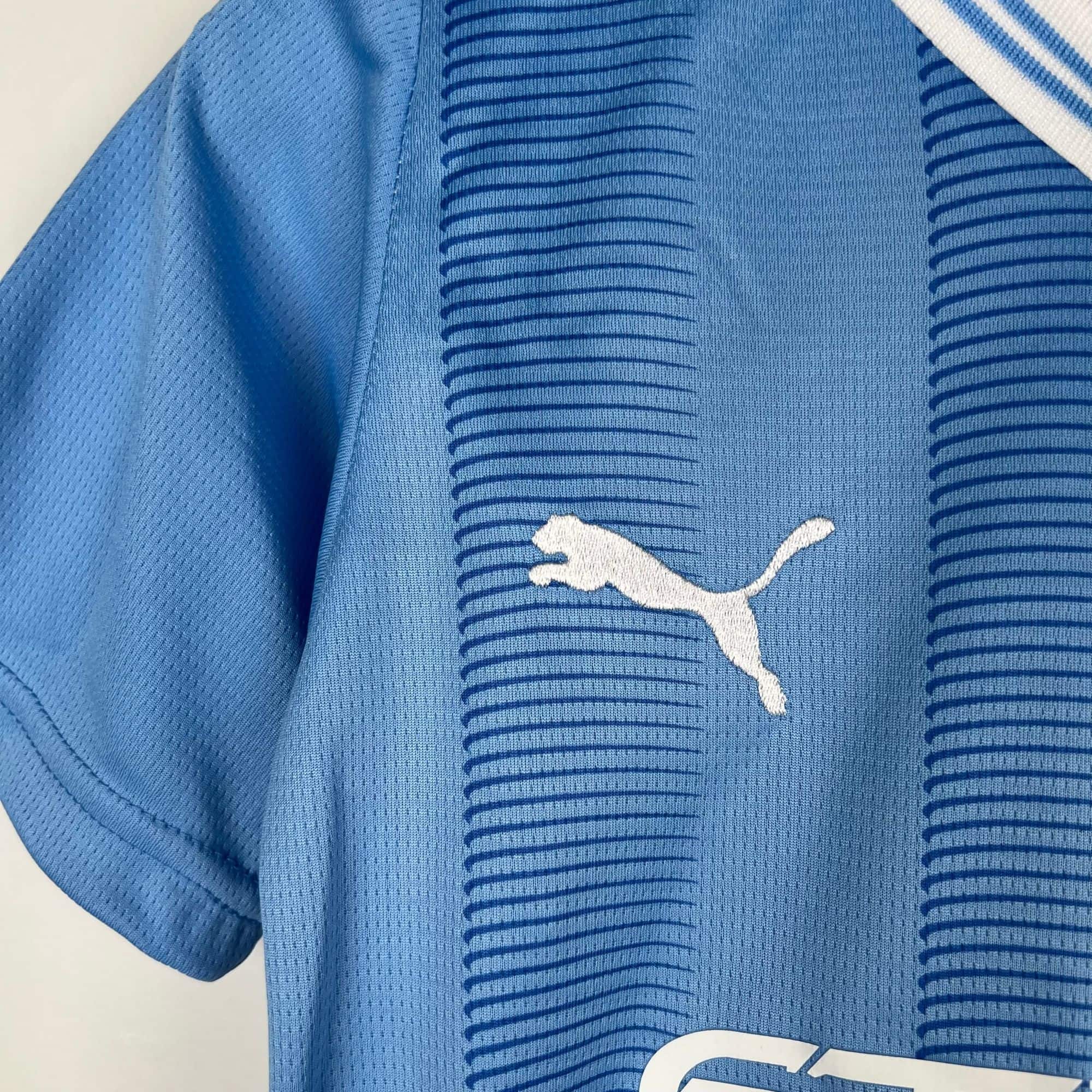 f8ed8e6e_b345af90-1501-45bb-aa63-432577cd1fa4 | UAE SPORT STORE Close-up of blue athletic jersey fabric with small perforations and a partial white logo/lettering visible at the bottom-right.