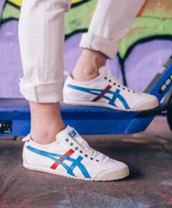 Close-up of white sneakers with blue and red stripes, rolled cuffs, standing on a blue scooter with a graffiti wall in the background.”] ,