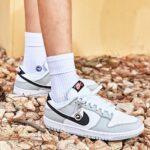 White and gray Nike sneakers with black swoosh on rocky ground, paired with white crew socks.
