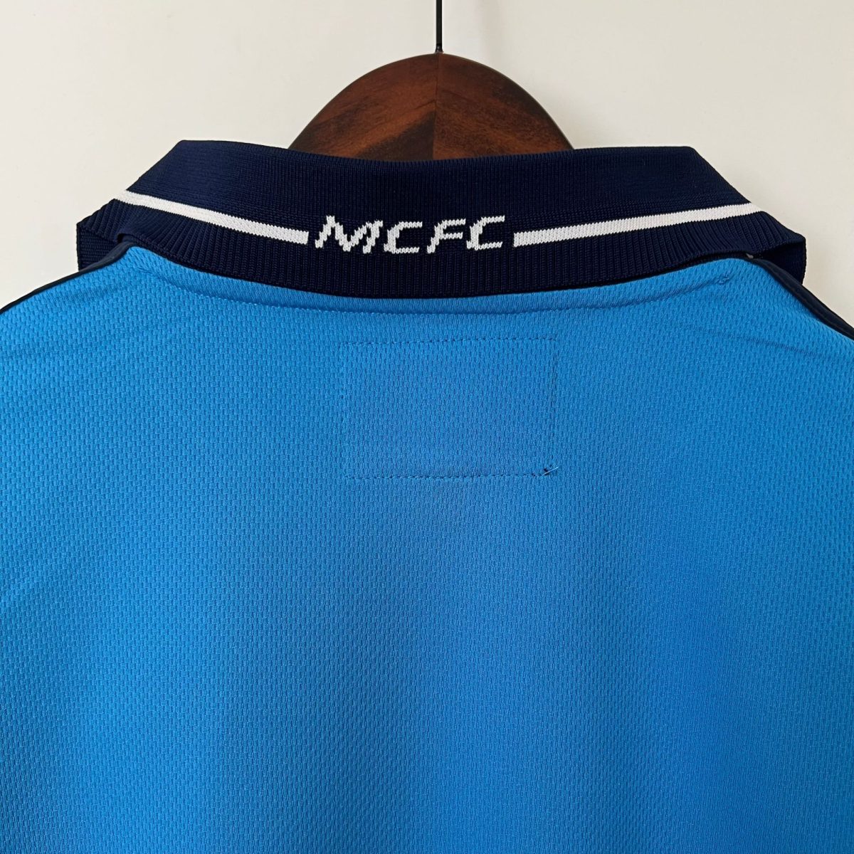 Blue polo shirt on a wooden hanger, navy collar with a white stripe and white embroidered script text on the back of the collar (text unreadable).