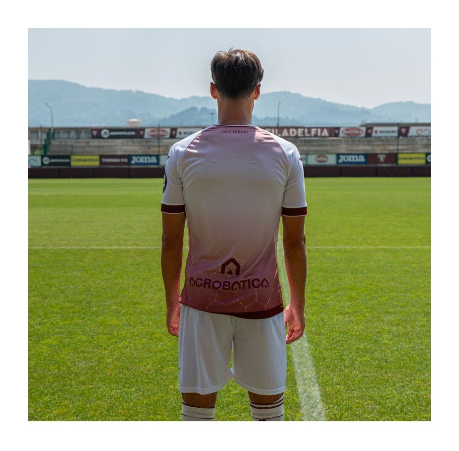 fCfEzzNzqxBbO6T | UAE SPORT STORE Back view of a soccer player standing on a sunny green pitch, wearing a pink-white gradient jersey and white shorts, with stadium banners in the background.