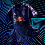 Navy blue soccer jersey with light blue wave pattern, Red Bull logo center, white shoulder stripes, and a small crest on the chest against a neon, wavy backdrop.