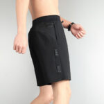 Male model wearing black Nike athletic shorts in a side view, showing a stitched panel design and a vertical Nike logo stripe along the leg seam.