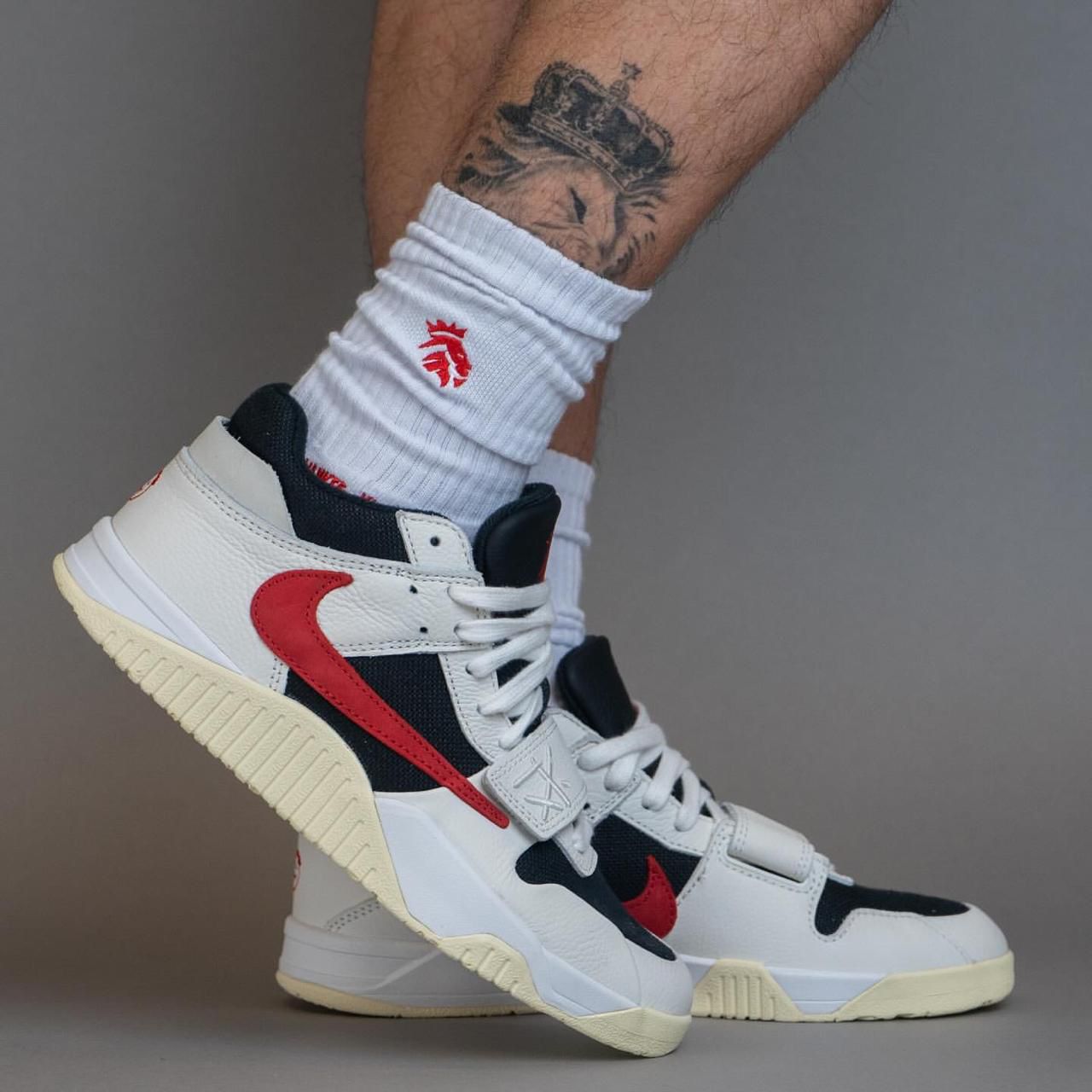 fa7999c7-859b-4846-aaad-a28b7d6aa47b | UAE SPORT STORE White high-top sneaker with red swoosh and navy panels worn with white ribbed sock; a tattooed leg is in the background.