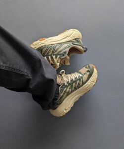 Person wearing green and beige HOKA trail shoes with a chunky beige sole, legs crossed on a blue floor.