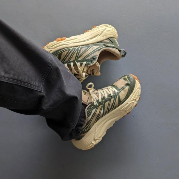 Person wearing green and beige HOKA trail shoes with a chunky beige sole, legs crossed on a blue floor.