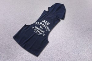 Navy sleeveless hooded athletic shirt on a beige carpet, with bold white text reading 'IRON PARADISE' and 'SANCTUARY OF SWEAT' on the front.