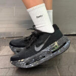 Black Nike running shoe with a gray marble-pattern midsole and white socks, on a paved surface.