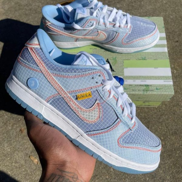 Close-up of light-blue grid-pattern Nike sneakers with pink stitching, white laces, and a yellow UN/LA tag, held in a hand over a green striped shoebox.