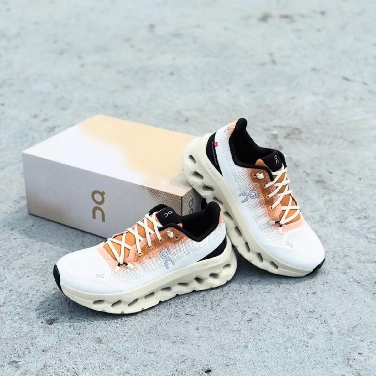 Pair of On running shoes with cream midsole and peach gradient upper beside a beige box.