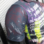 Close-up of a dark, graphic T-shirt with white repeated text and multicolor paint-splatter patterns on the sleeve and back.
