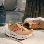 Beige running shoes with red 'ON' logo resting on a concrete path beside a tree, laces untied near a water bottle.