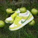 Bright lime-green and white sneakers with neon laces resting on grass among green apples nearby.
