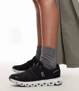 Side view of lower legs wearing black On running shoes with white soles and gray socks, beige pants showing at the top edge