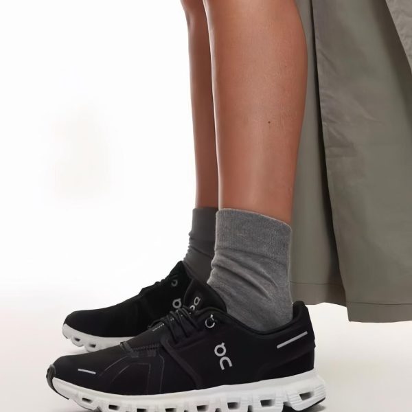 Side view of lower legs wearing black On running shoes with white soles and gray socks, beige pants showing at the top edge