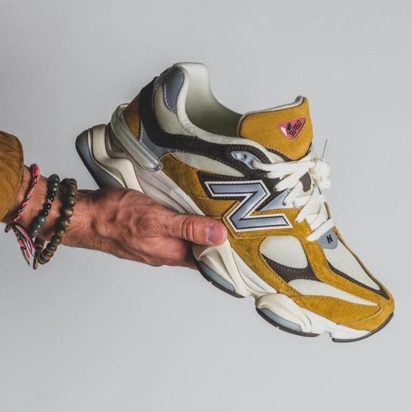 Person's hand with beaded bracelets holding a mustard-yellow and white New Balance sneaker against a plain gray background, side view.